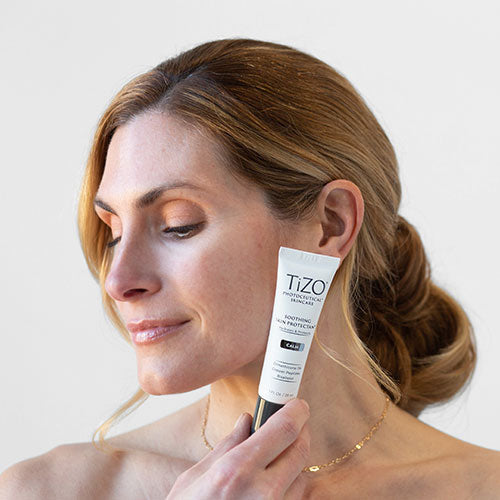Model from shoulders up with brown hair holding TiZO Soothing Skin Protection bottle against cheek