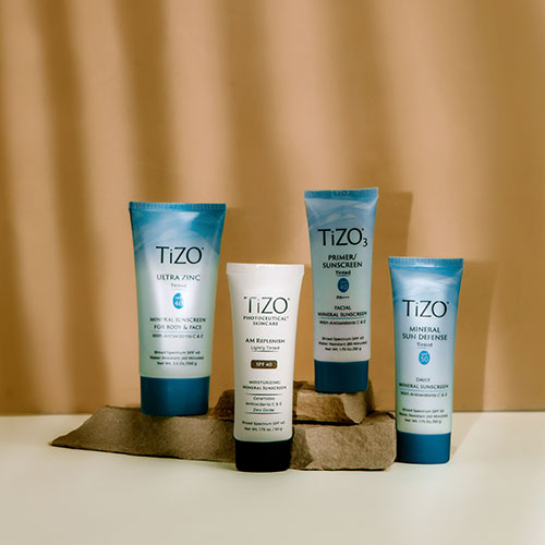 Four TiZO tinted mineral sunscreen products lined up on rocks with a tan background and light shining down